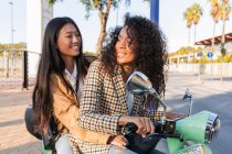 Mujer asiática optimista con sonrisa dentada y mujer afroamericana satisfecha con el pelo rizado mirándose mientras está sentada en la moto - foto de stock