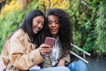 Pleasant Asian female with toothy smile showing video on smartphone to happy black woman while spending time together — Fotografia de Stock