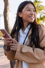 Felice donna asiatica in abito elegante in piedi sulla strada e navigazione internet sul telefono cellulare — Foto stock