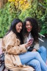Pleasant Asian female with toothy smile showing video on smartphone to happy black woman while spending time together — Photo de stock