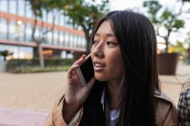 Asian female in stylish outfit standing on street and talking phone — Fotografia de Stock