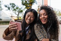 Asiatin hat Spaß am Telefonieren auf dem Smartphone und fokussierte schwarze Frau hört Musik und surft mit dem Handy im Internet — Stockfoto