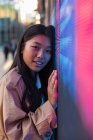 Wistful ethnic female in casual clothes with long hair looking at camera while leaning on wall with neon illumination — Fotografia de Stock