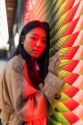Wistful ethnic female in casual clothes with long hair looking at camera while leaning on wall with neon illumination — Fotografia de Stock