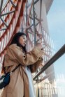 From below of serious asian woman in stylish outfit looking out window while taking picture on mobile phone — Fotografia de Stock