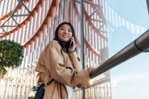 Вид збоку щасливої азіатки в стильному вбранні, дивлячись далеко під час розмови на мобільному телефоні — стокове фото