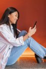 Glückliche asiatische Frau in lässiger Kleidung sitzt in bequemer Pose und surft auf dem Handy im Internet — Stockfoto