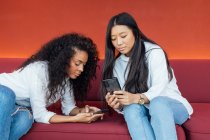 Young black female and satisfied Asian girlfriend sitting on sofa and browsing smartphone while resting — Stock Photo
