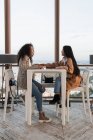 Seitenansicht von stilvollen enthusiastischen multiethnischen Freundinnen, die sich unterhalten und am Tisch in der Nähe des Fensters im Café sitzen — Stockfoto