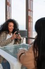 Mujer negra tomando fotos de mujer asiática sentada a la mesa en la cafetería ligera - foto de stock