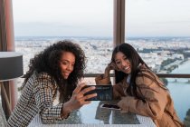 Junge fröhliche schwarze Frau und zufriedene asiatische Freundin sitzt auf dem Sofa und macht ein Selfie auf dem Handy, während sie sich ausruht — Stockfoto