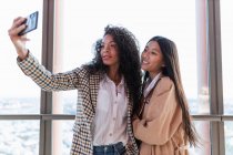 Junge fröhliche schwarze Frau und zufriedene asiatische Freundin steht im Café und macht Selfie auf dem Handy — Stockfoto