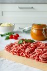 From above of fresh raw mixed pork and beef minced meat on wooden cutting board with green basil leaves placed on table during cooking process near homemade tomato sauce — Fotografia de Stock