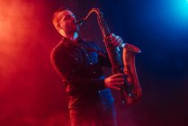 Professional male musician with eyes closed playing saxophone in red and blue neon lights during live performance — Stock Photo