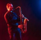 Professional male musician with eyes closed playing saxophone in red and blue neon lights during live performance — Fotografia de Stock