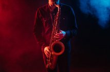 Músico de la cosecha tocando saxofón en luces de neón rojas y azules durante la actuación en vivo - foto de stock