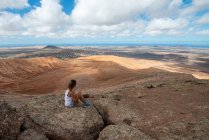 Side view female hiker in casual outfit sitting on rocky hill and enjoying spacious hilly valley on clear day — Photo de stock