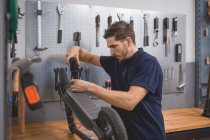 Seitenansicht des männlichen Mechanikers mit Schraubenzieher, der das Rad des Elektrorollers in der Werkstatt fixiert — Stockfoto