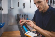 Side view of concentrated skilled male mechanic using multimeter while testing battery of electric scooter in workshop — Stock Photo