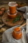 Full glass jars with homemade natural fried tomato sauce placed on cutting board board near with fresh green rosemary and basil leaves placed on rustic wooden table — Stock Photo