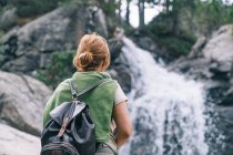Back view of anonymous female hiker in casual clothes with backpack standing in forest admiring views of powerful waterfall — Stock Photo