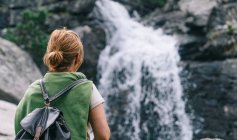 Back view of anonymous female hiker in casual clothes with backpack standing in forest admiring views of powerful waterfall — Stock Photo