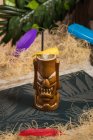 Tasse à tiki sculpturale marron avec boisson alcoolisée décorée de paille et de glace placée au milieu de l'herbe sèche contre une plante verte avec plume sur fond flou — Photo de stock
