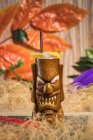 Brown sculptural tiki mug with alcohol drink decorated with straw and ice placed amidst dry grass against green plant with feather on blurred background — Stock Photo