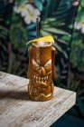 Tazza di tiki scultorea marrone con bevanda alcolica decorata con paglia e ghiaccio posta sul tavolo su sfondo sfocato — Foto stock