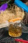 Taza de vidrio Tiki con bebida antigua colocada sobre tela en medio de hierba seca contra valla de madera y hojas coloridas sobre fondo borroso - foto de stock