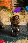 Керамическая полинезийская кружка с алкогольным напитком подается с соломой и украшения помещены на ткань против сухой травы и красочные листья — стоковое фото