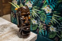 Tasse à tiki sculpturale marron avec boisson alcoolisée décorée de paille et de glace placée sur une table en bois — Photo de stock