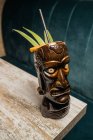 Brauner skulpturaler Tiki-Becher mit Alkoholgetränk verziert mit Stroh und Eis auf Holztisch — Stockfoto