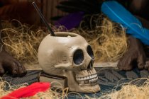 Ceramic polynesian tiki cup skull shaped with straw placed amidst dry grass with wooden fence and colorful feathers on blurred background — Stock Photo