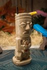 Traditionelle skulpturale Tiki-Tasse mit Alkohol und Stroh auf Teppich gegen Holzzaun gelegt bunte Blätter und trockenes Gras — Stockfoto