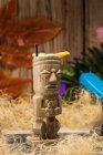 Copo tradicional escultural tiki de bebida alcoólica com palha colocada no tapete contra cerca de madeira folhas coloridas e grama seca — Fotografia de Stock