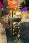 Сверху большая скульптурная чашка с тиками, наполненная выпивкой, украшенной соломой и фруктами, помещенными на зеленом ковре против сухой травы — стоковое фото