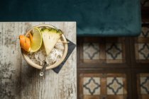 De cima de grande copo de tiki escultural cheio de bebida decorada com palha e frutas colocadas no tapete verde contra mesa de madeira — Fotografia de Stock
