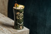 Grande copo de tiki escultural cheio de bebida decorada com palha e frutas colocadas no tapete verde contra mesa de madeira — Fotografia de Stock