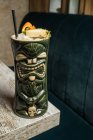 Большая скульптурная чашка с тиками, наполненная выпивкой, украшенной соломой и фруктами, помещенными на зеленом ковре против деревянного стола — стоковое фото