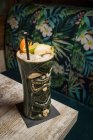 Grande tasse sculpturale tiki remplie d'alcool décoré de paille et de fruits placés sur un tapis vert contre une table en bois — Photo de stock