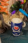 De dessus de tiki en forme de taureau tasse de boisson alcoolisée avec mousse placée contre l'herbe sèche et les plumes sur fond flou — Photo de stock