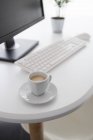 Computer moderno con monitor nero e tastiera bianca posizionato sulla scrivania con pianta verde in vaso e tazza di caffè in ufficio — Foto stock