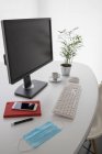 Moderner Computer und Smartphone mit Notizbuch auf weißem Tisch mit medizinischer Maske und Topfpflanze im hellen Büro — Stockfoto