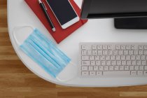 Современный компьютер и смартфон с ноутбуком помещен на белый стол с медицинской маской в светлом офисе — стоковое фото