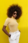 Молодая женщина с афропрической и солнцезащитными очками, стоящая на желтой стене — стоковое фото