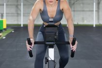 Determined muscular female athlete in sportswear riding stationary bicycle during functional workout in sport club — Stock Photo