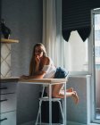 Привлекательная мечтательная женщина в белом топе с голыми плечами, держащими сладкий персик и смотрящая в камеру, опираясь на кухонный стол — стоковое фото