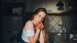 Приваблива мрійлива жінка в білому верху з голими плечима, що тримає солодкий персик і дивиться на камеру, спираючись на кухонну стійку — стокове фото