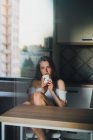 Gelassene junge Frau mit langen Haaren trägt lässige Kleidung mit nackten Schultern, genießt frisches Heißgetränk und blickt in die Kamera, während sie sich auf den Küchentisch lehnt — Stockfoto
